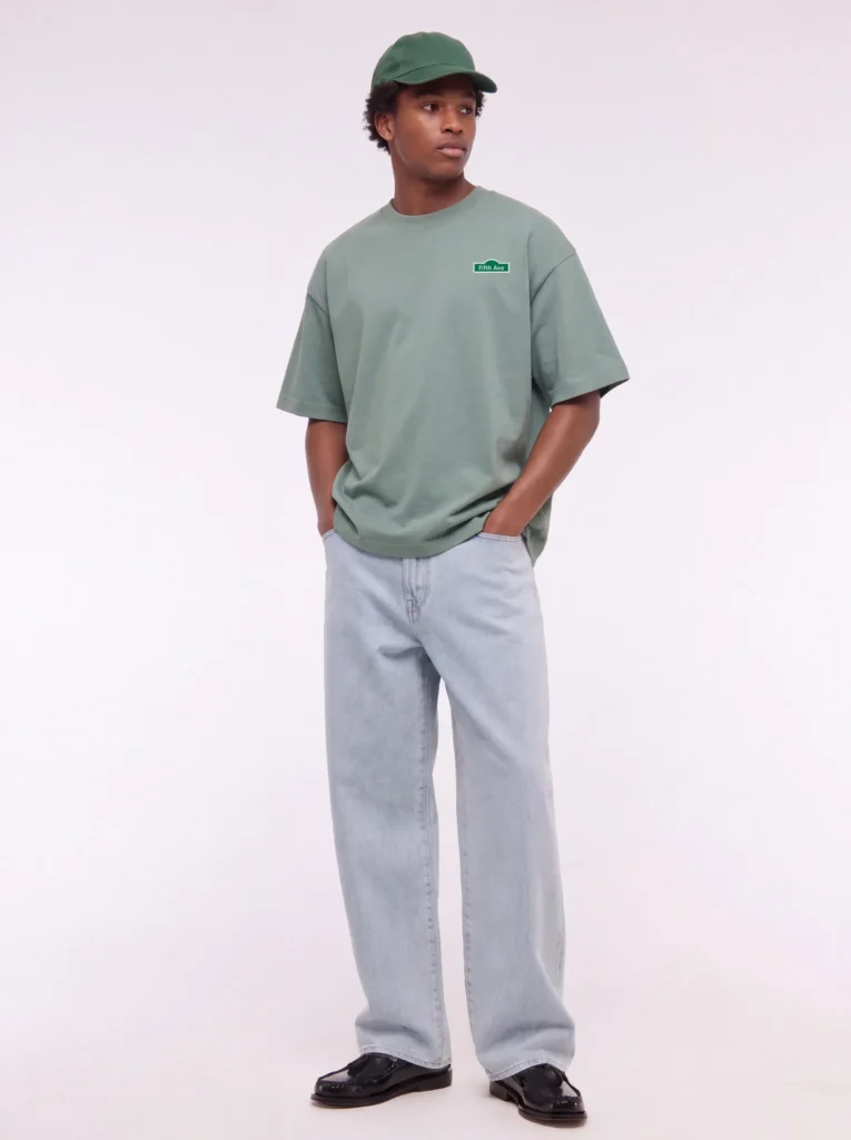 Model in a sage-green NeedleWorks heavyweight T-shirt with an embroidered patch, green cap and light-wash jeans